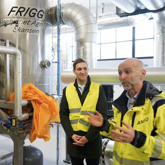 Two men standing in front of an industrial heat pump that says "Frigg" on it